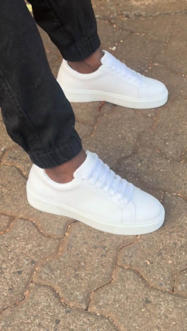 White leather low-top sneakers paired with black trousers on a cobblestone path.