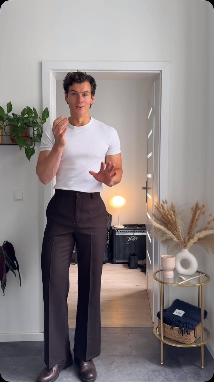 Man wearing a white t-shirt and dark brown trousers standing in a doorway