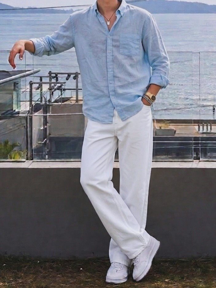 Man wearing a light blue shirt and white trousers standing by the ocean.