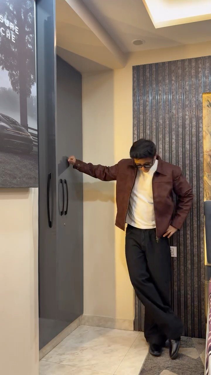 Man wearing a brown jacket, white t-shirt, and black trousers standing indoors.