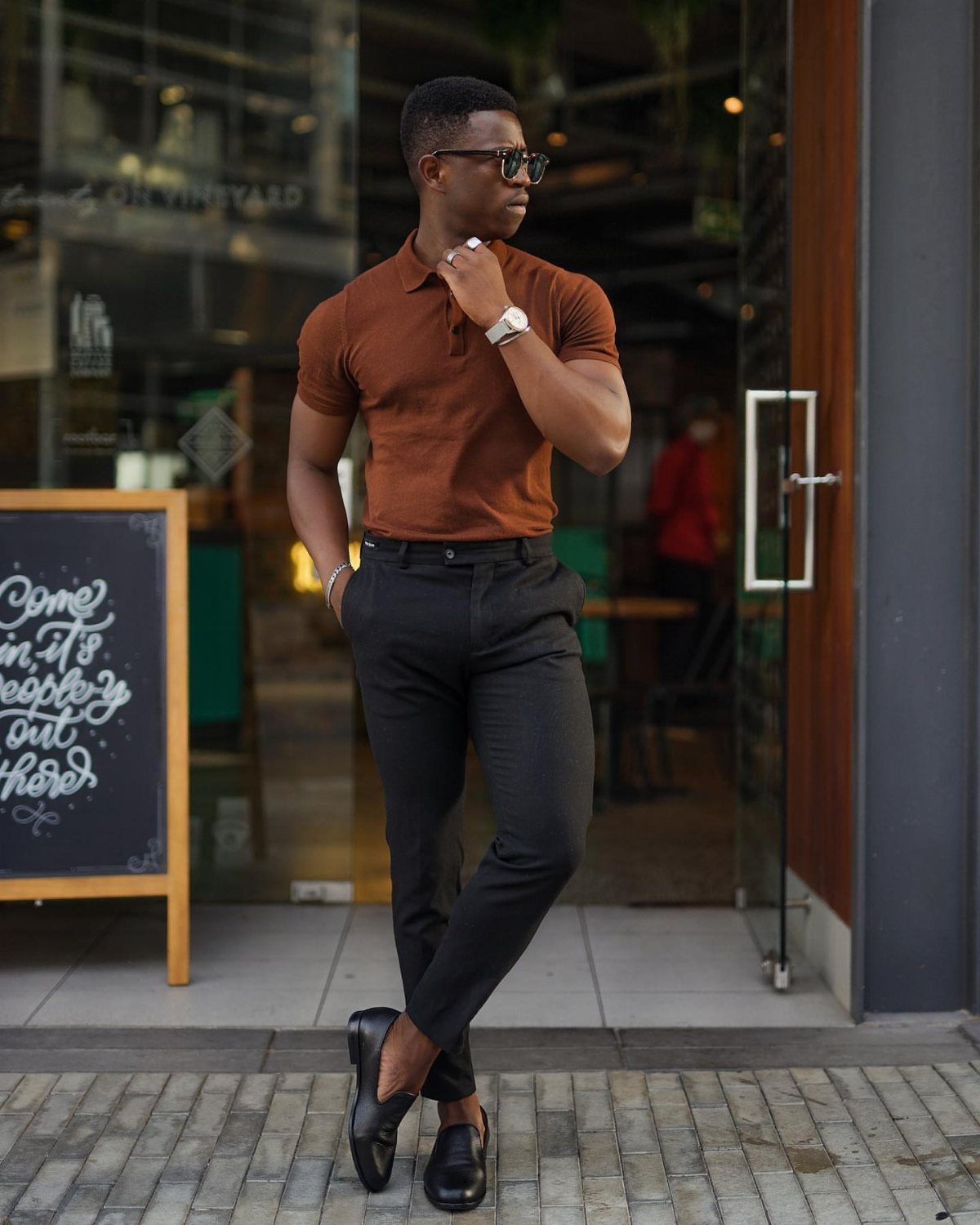 Man in brown polo and black trousers wearing black leather loafers outdoors.