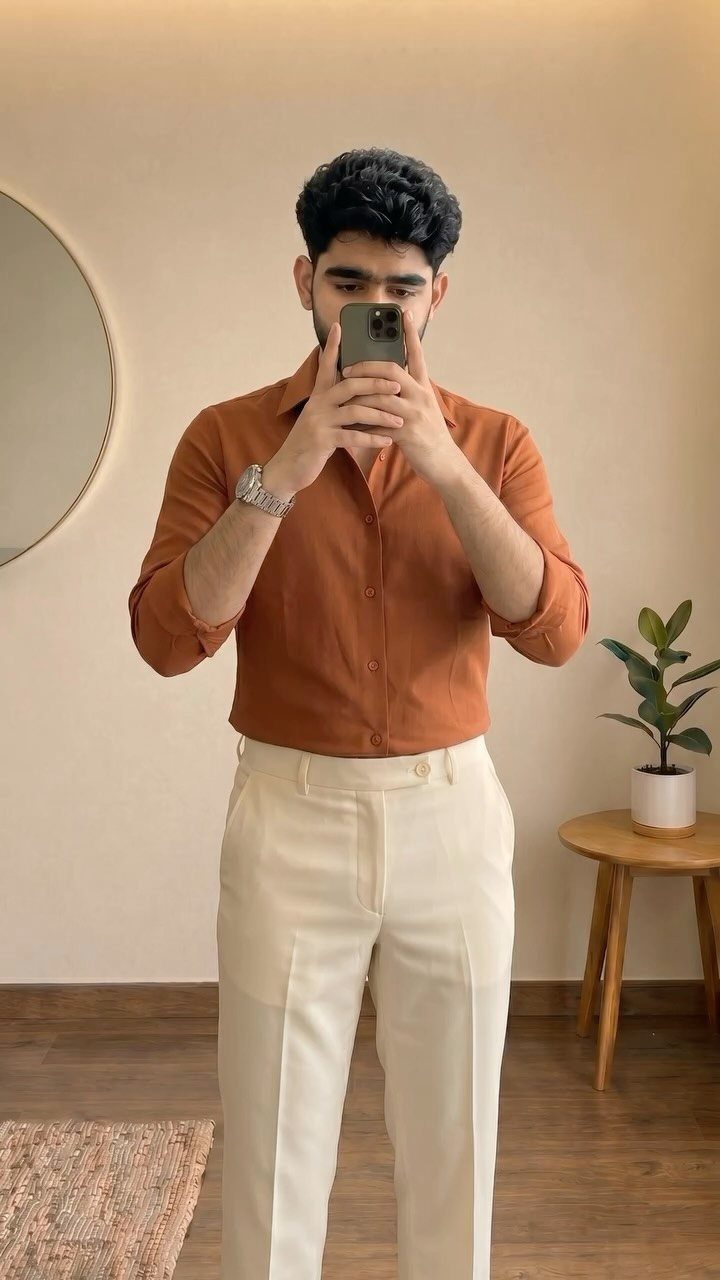 Man in rust-colored shirt and off-white trousers taking a mirror selfie indoors.