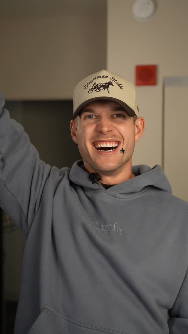 Smiling man wearing a grey hoodie and a tan baseball cap indoors.