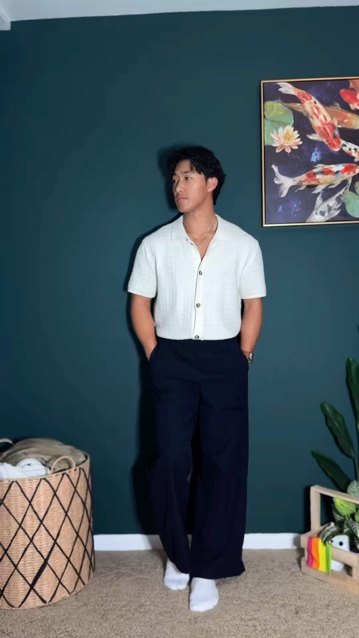 Man wearing a white short-sleeved shirt and wide-leg navy trousers against a teal wall