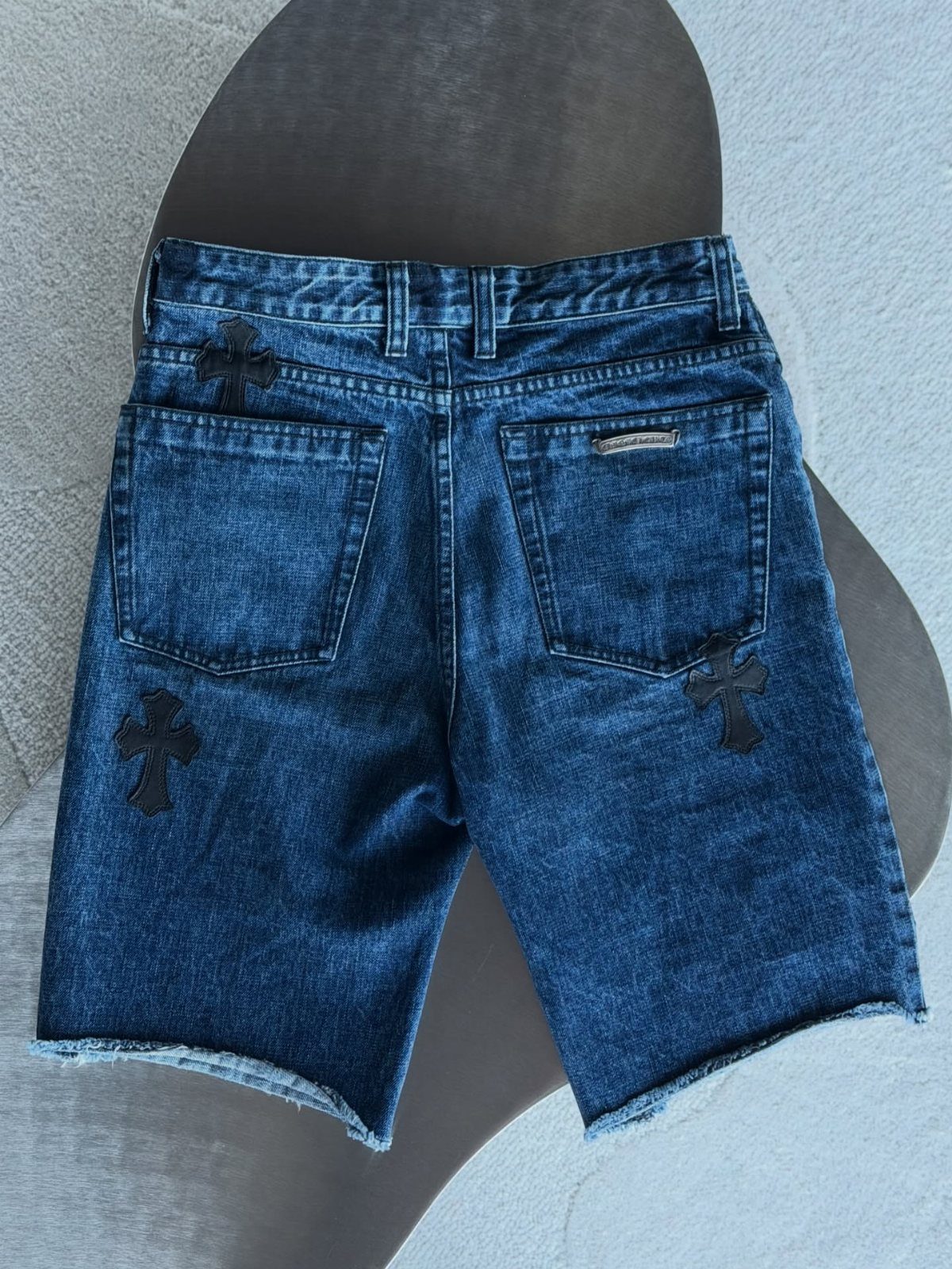 Dark blue denim jean shorts with black cross patches on the back pockets.