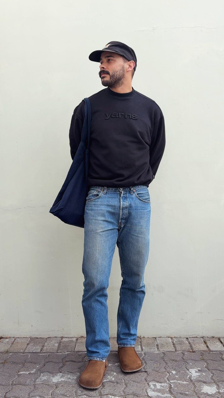 Man wearing a black sweatshirt, blue jeans, and brown shoes against a white wall.