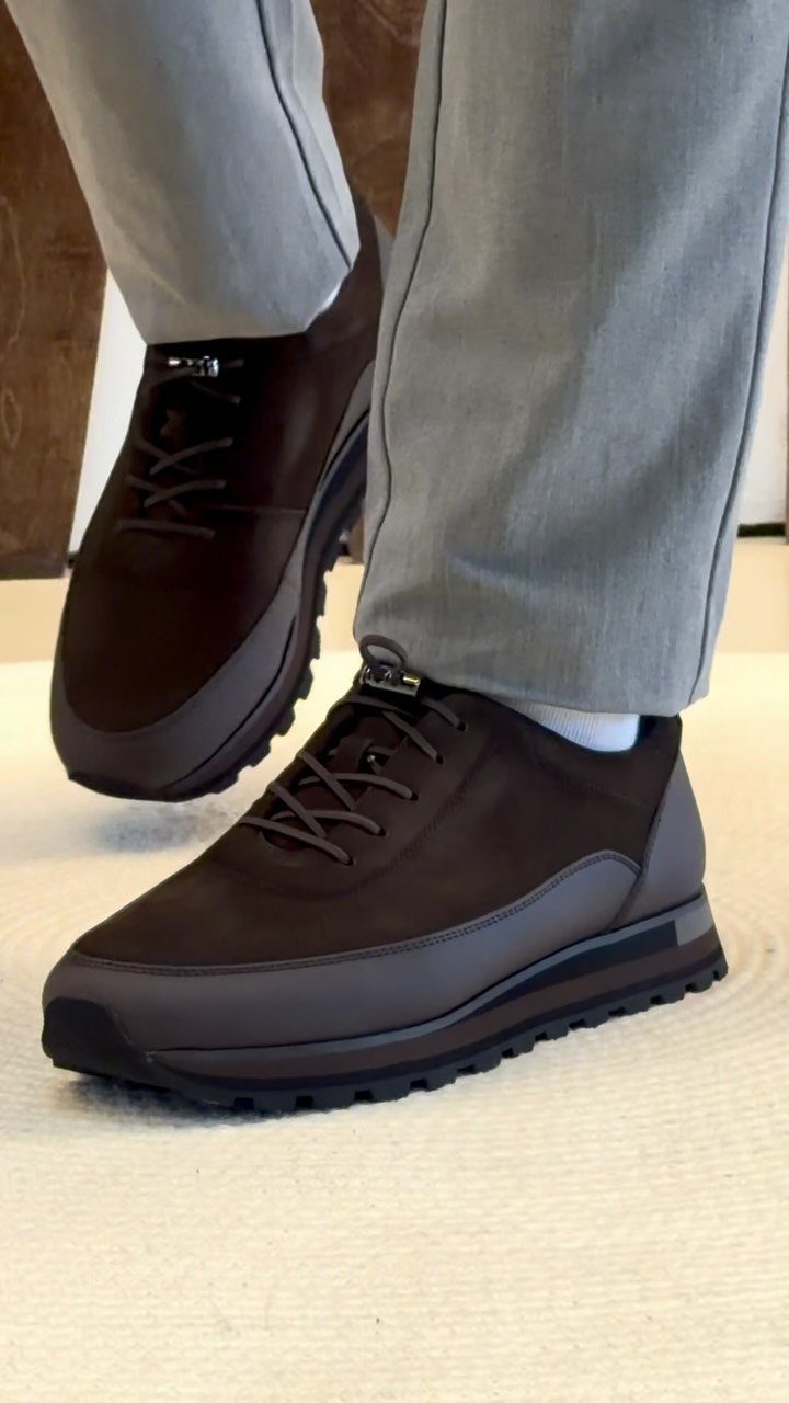 Dark brown minimalist sneakers paired with grey tailored trousers on a light floor.