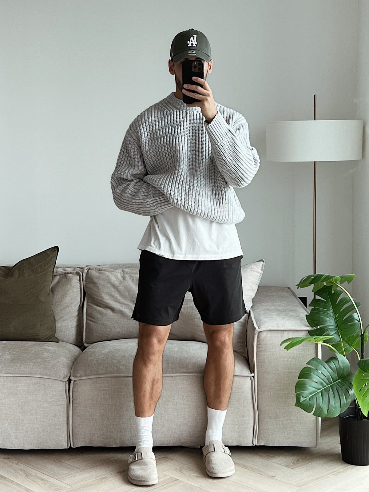 Man wearing oversized grey sweater, black shorts, and beige clogs in a living room.