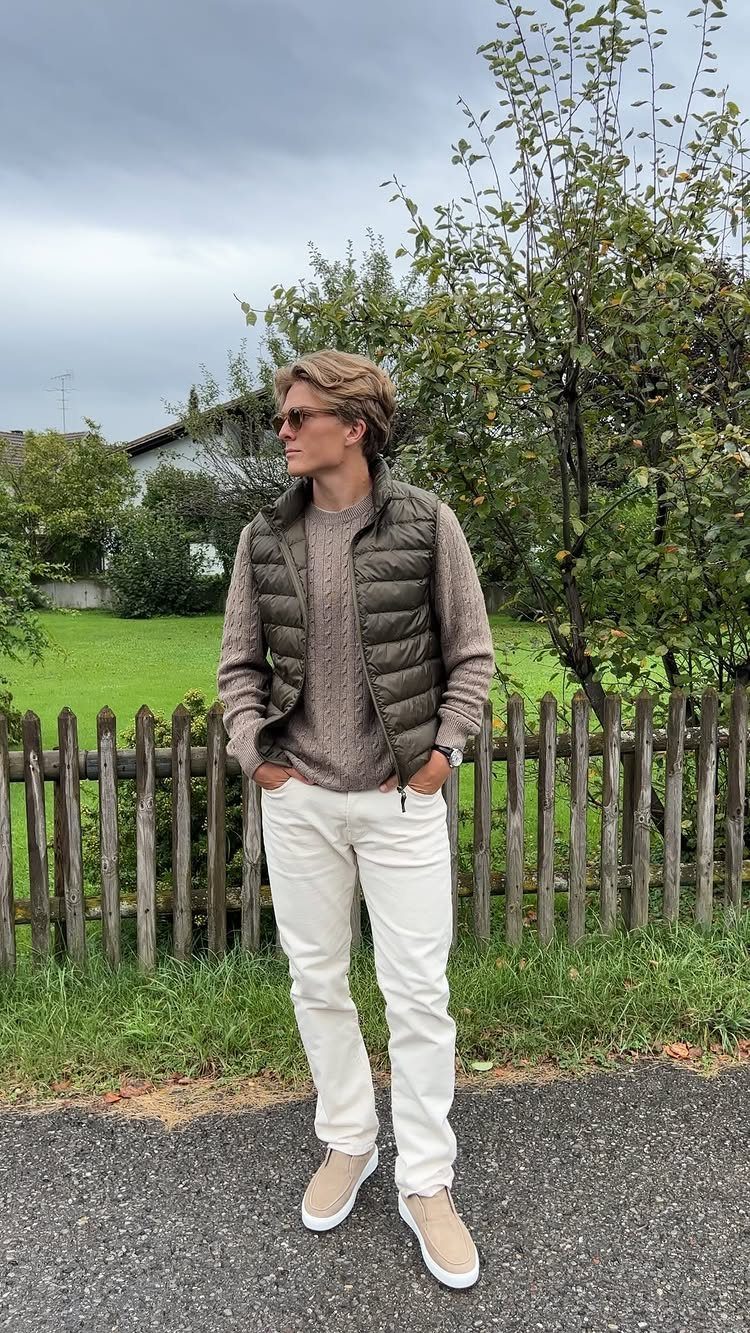 Man wearing a brown puffer vest, beige sweater, and white trousers outdoors.