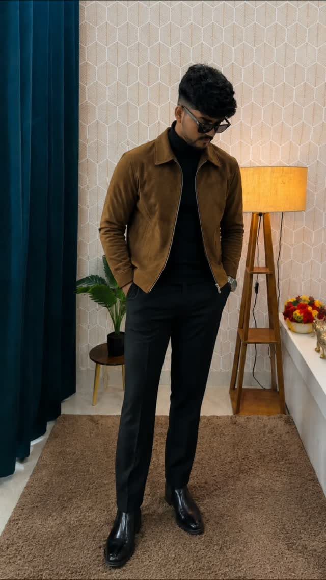 Man in brown suede jacket, black turtleneck, and black trousers posing indoors.