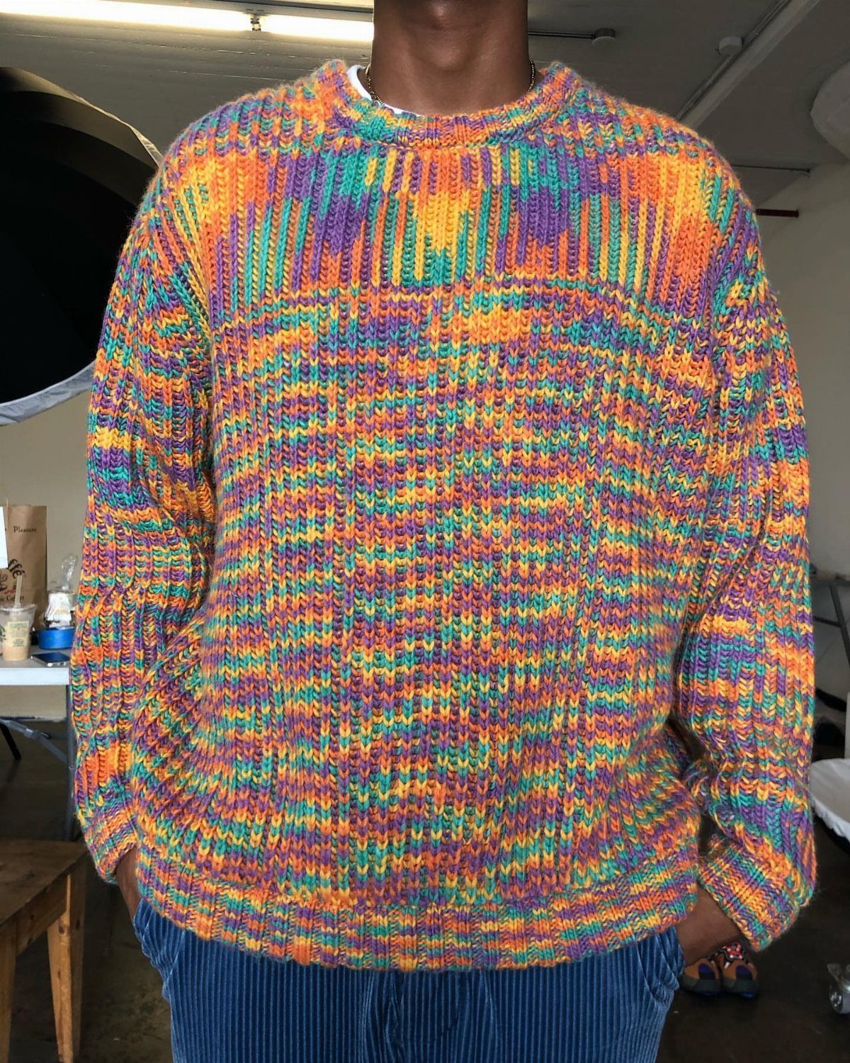 Person wearing a rainbow multi-colored knit sweater and blue corduroy pants indoors.
