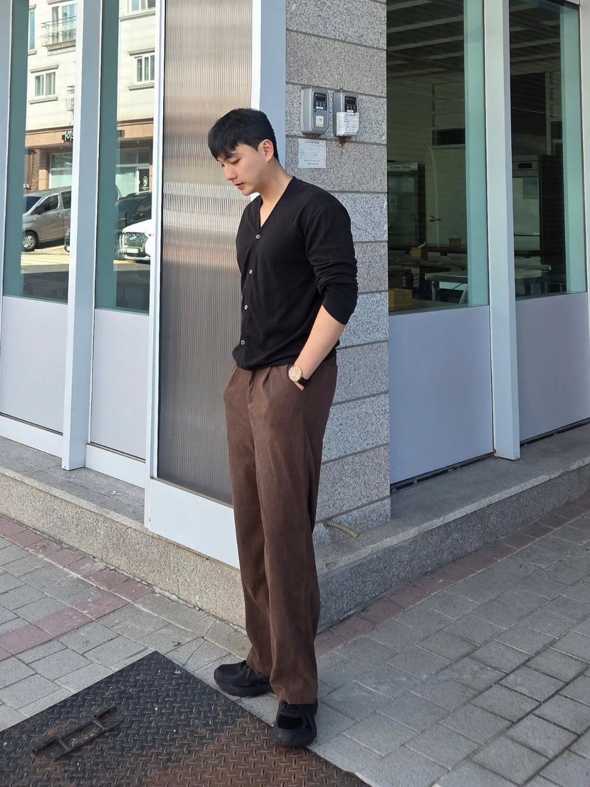 Man wearing a black shirt and brown corduroy trousers standing by a building.