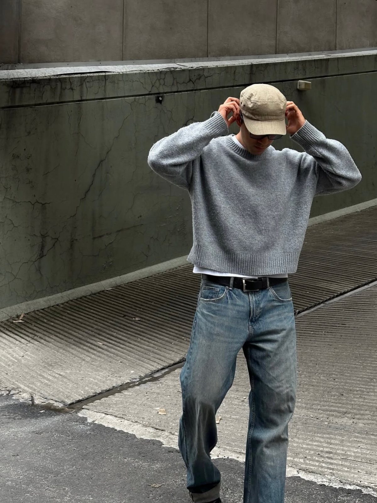 Man wearing a grey cropped knit sweater and blue jeans with a tan cap.