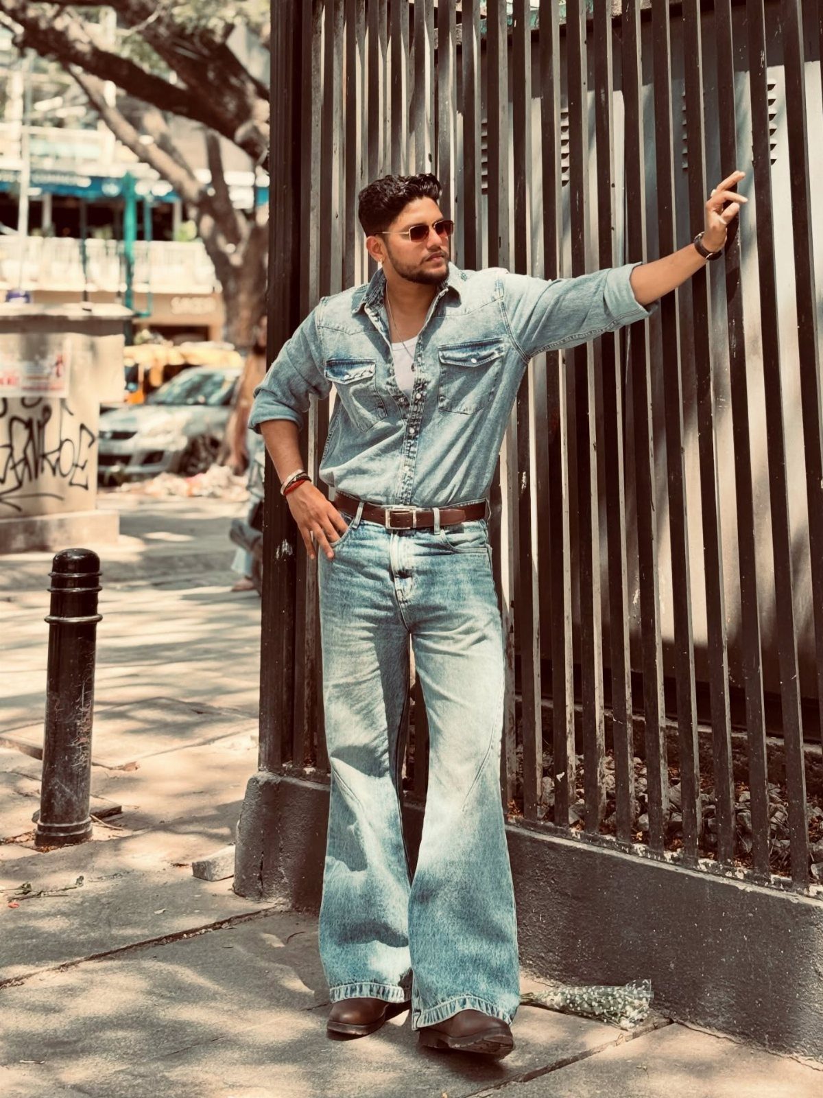 Man in light blue denim shirt and flared jeans posing by metal fence