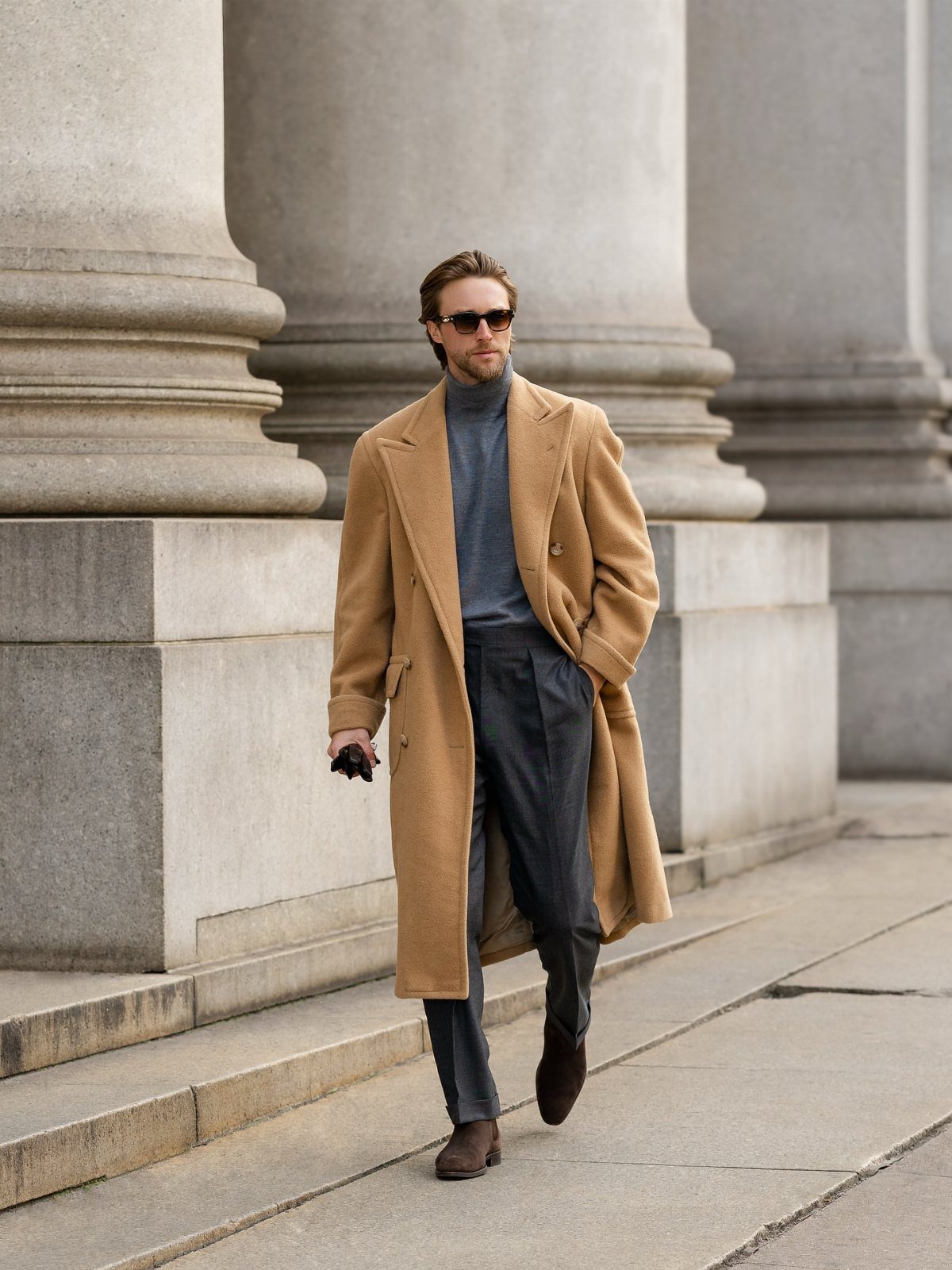 Man in a long camel-brown overcoat and grey turtleneck walking by stone columns.