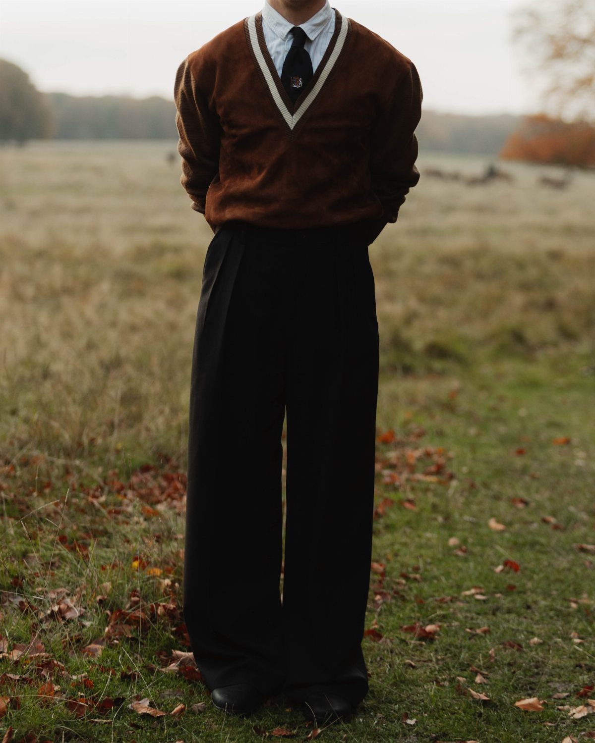 Person wearing a brown V-neck sweater and black trousers in an autumn field.