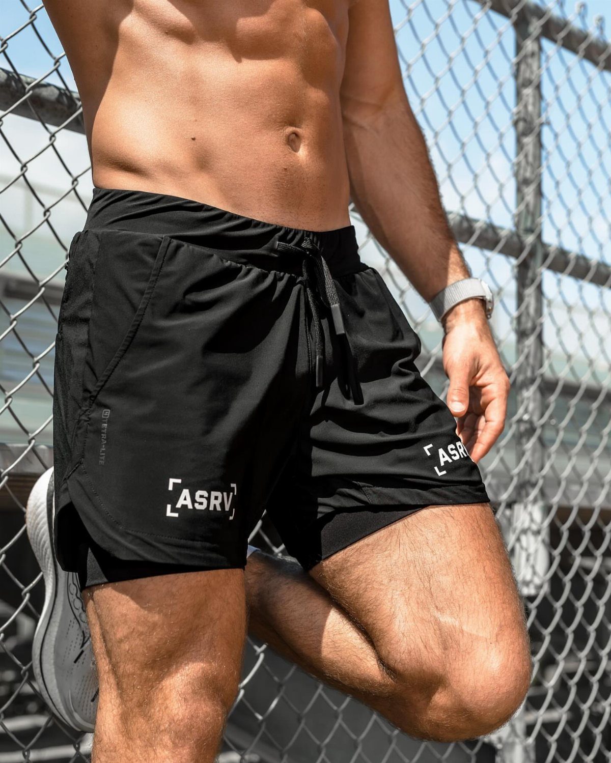 Shirtless man wearing black athletic gym shorts and white sneakers by a fence
