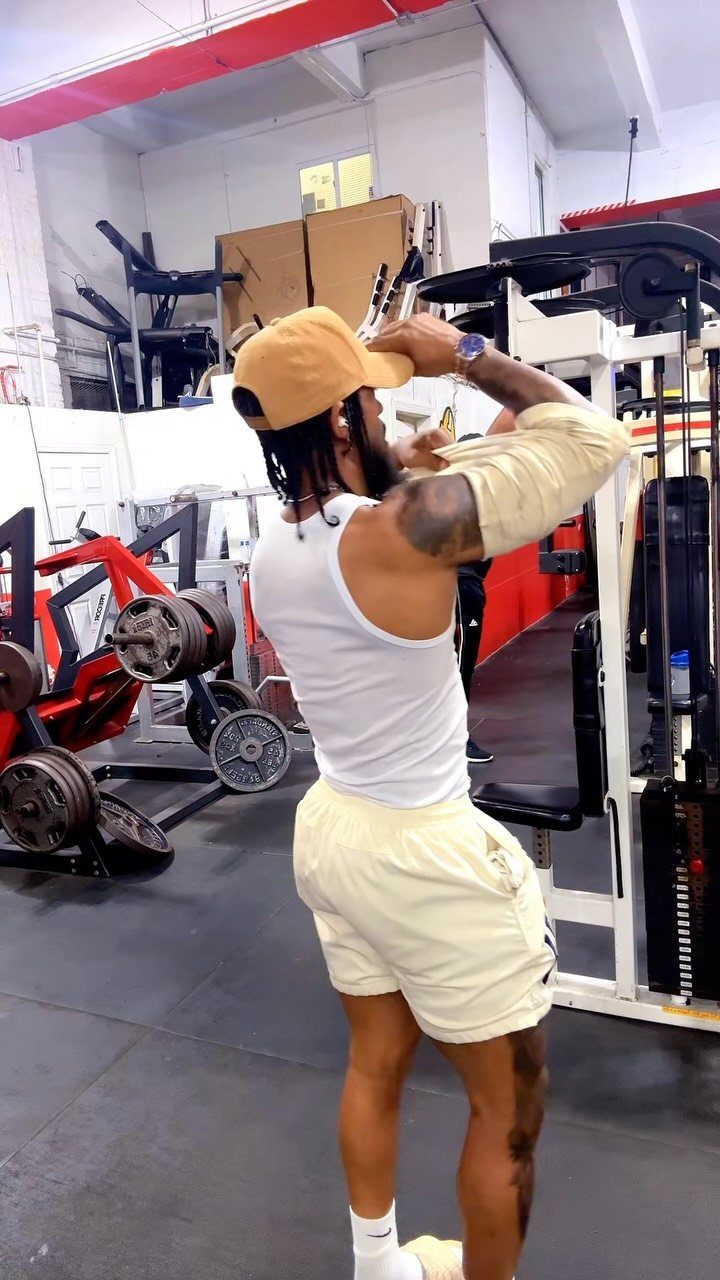 Man in white tank top and cream shorts standing in a gym setting.