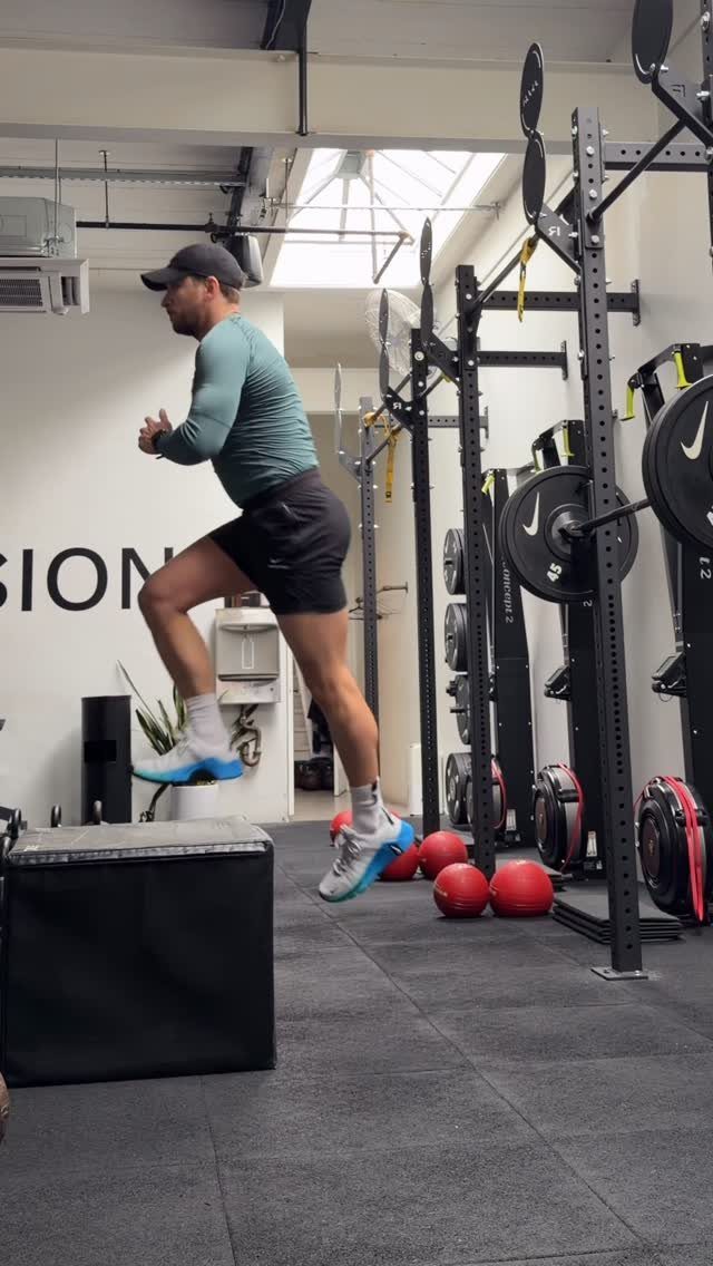 Man in teal athletic shirt and black shorts jumping over a gym box