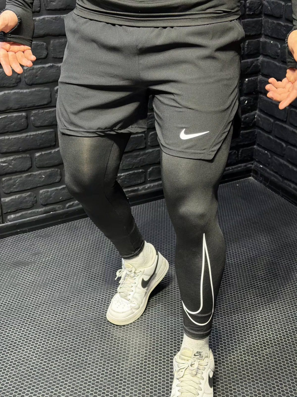 Man wearing black Nike shorts, black leggings, and white sneakers in a gym.