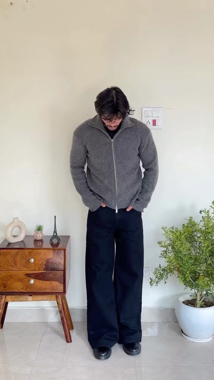Man wearing a gray zip-up cardigan and wide-leg black trousers standing indoors.