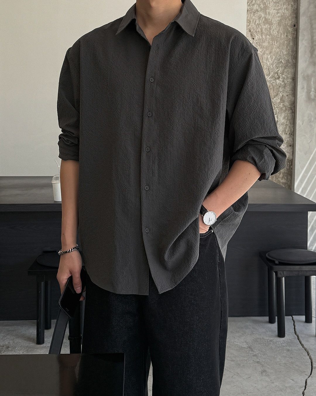 Person wearing a dark gray button-down shirt and black trousers in a minimalist room.