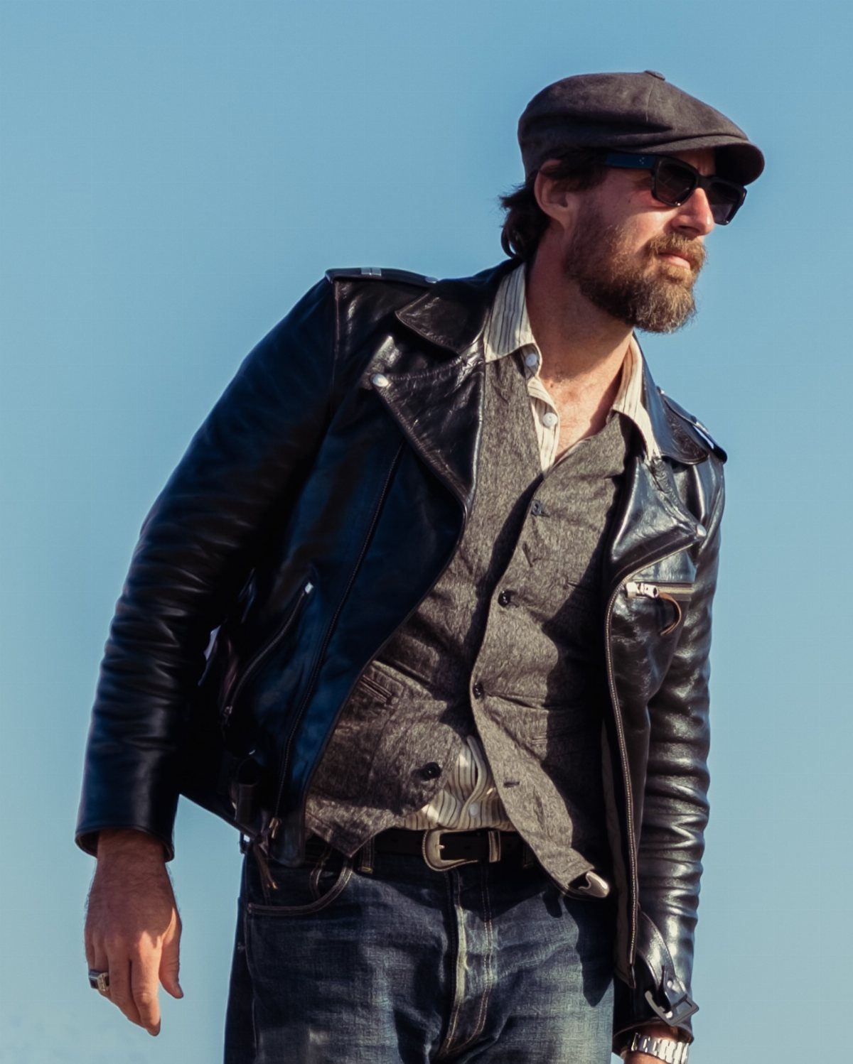 Man wearing a gray shirt, black leather jacket, and a dark flat cap.