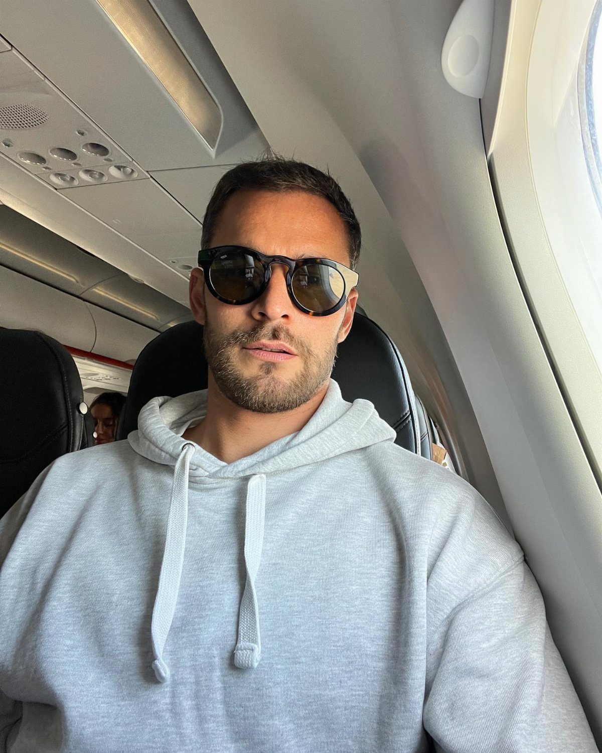 Man wearing sunglasses and a grey hoodie sitting inside an airplane cabin.