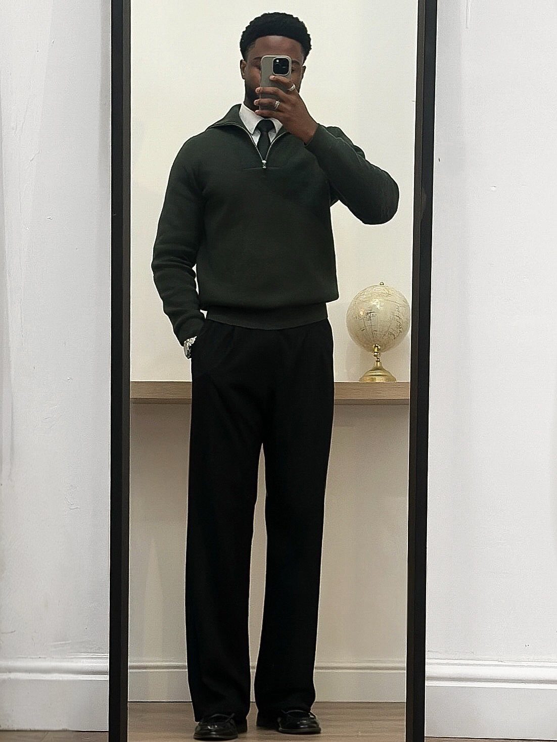 Man in dark green sweater and wide black trousers taking a mirror selfie