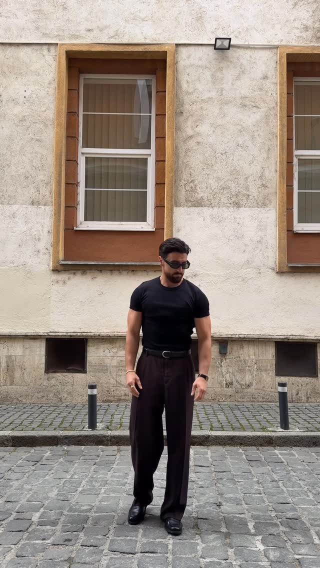 Man wearing a black t-shirt and brown wide-leg trousers on a cobblestone street.