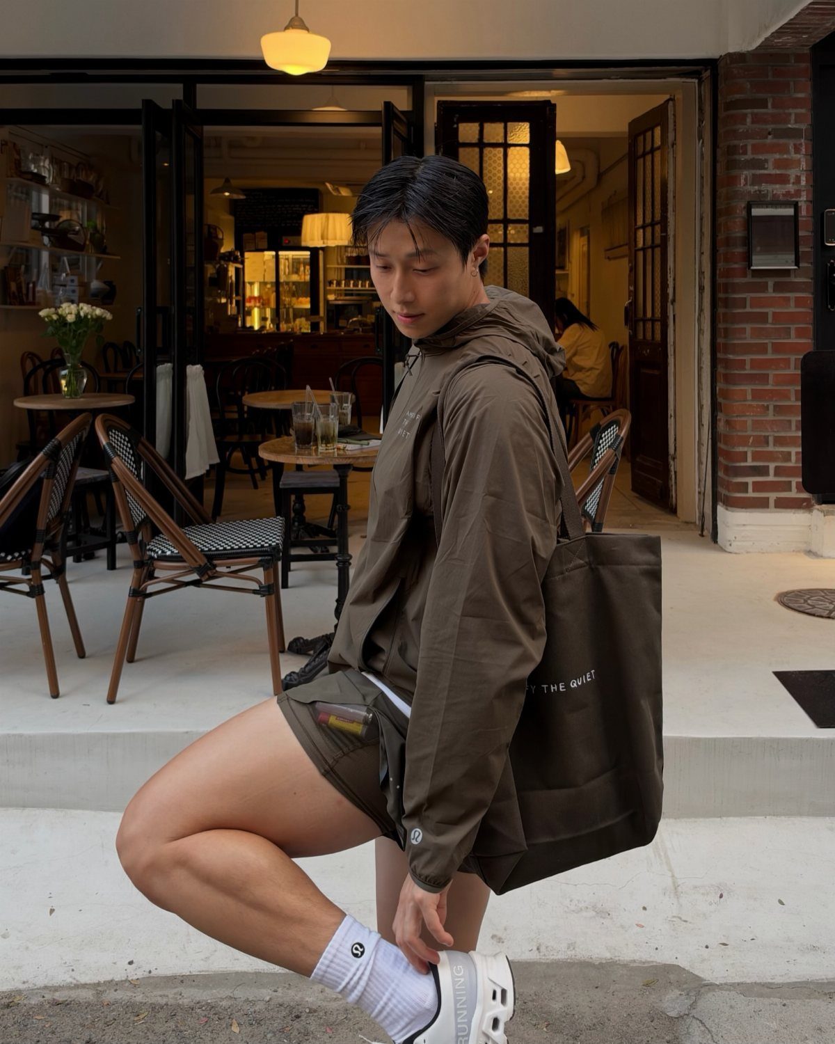 Man in olive green workout jacket and shorts posing outside a cafe.