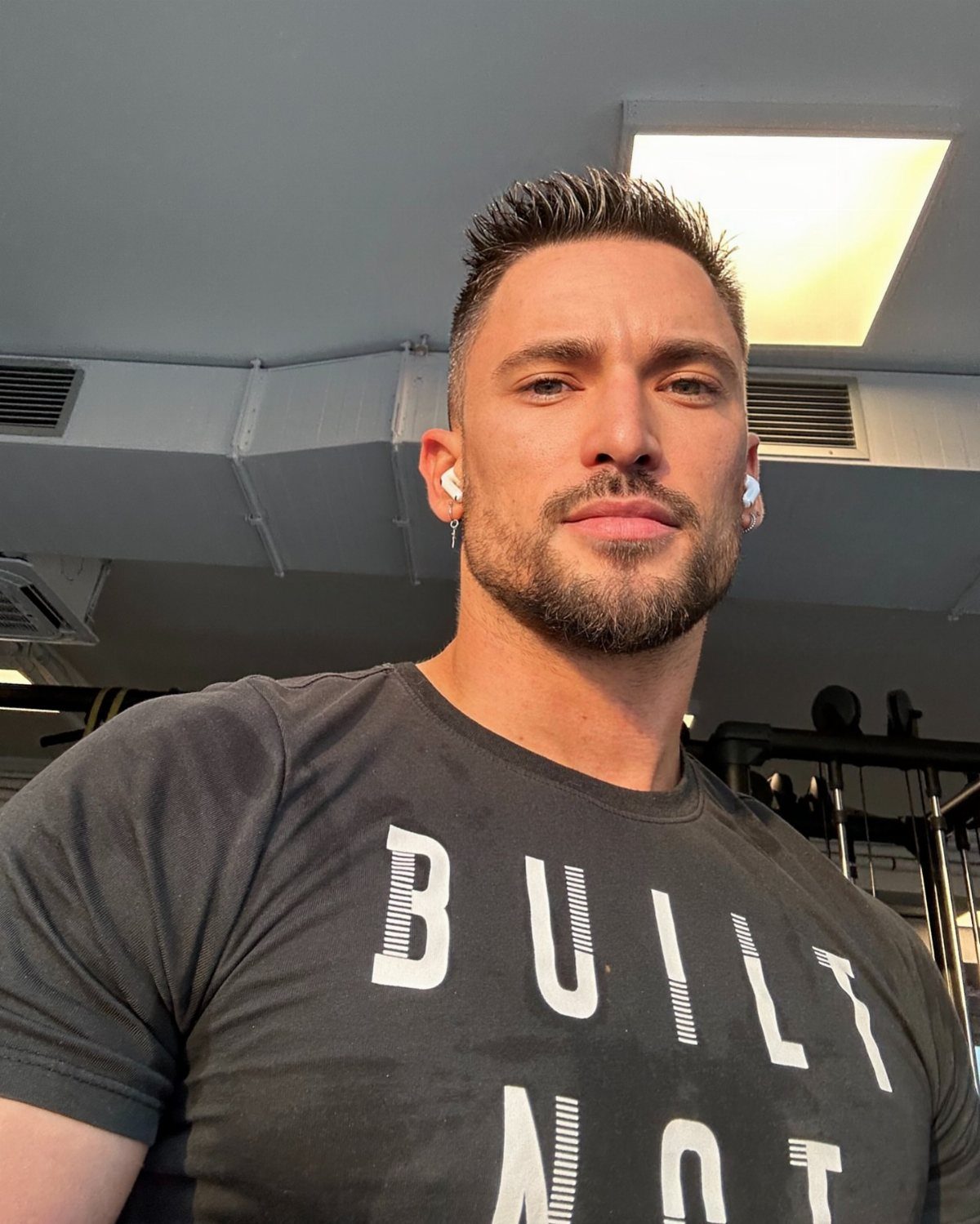 Muscular man in a black t-shirt posing for a selfie in a gym.