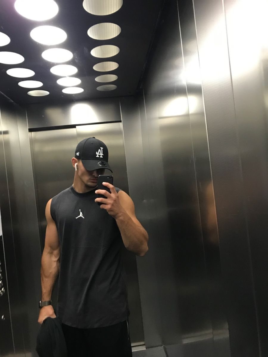 Muscular man taking a mirror selfie in an elevator with bright overhead lighting.