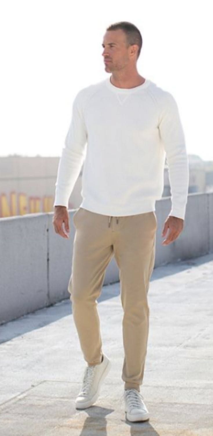 Man wearing a white sweater, tan joggers, and white sneakers walking outdoors.