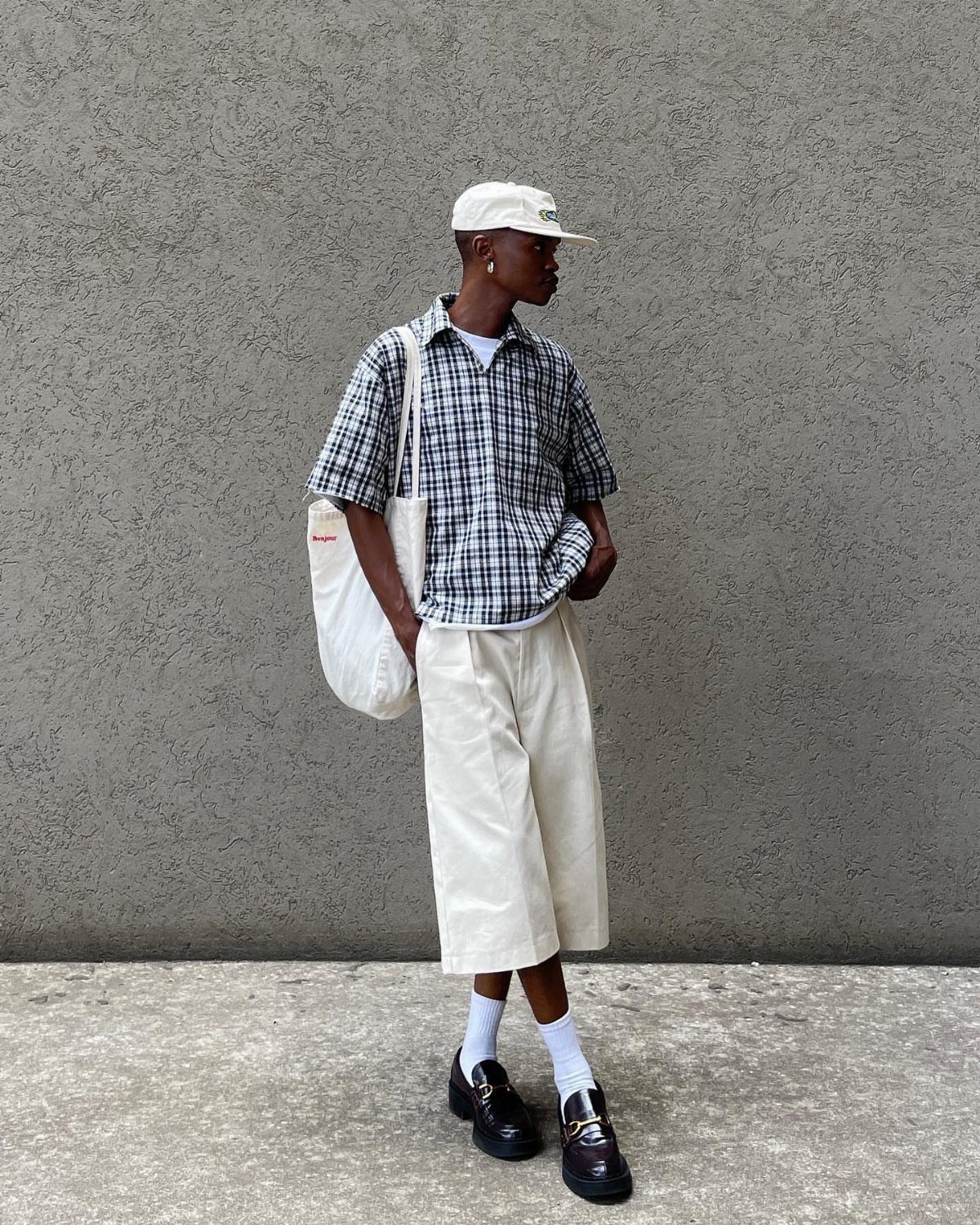 Man in checkered button-down shirt, white shorts, and black loafers standing by wall.
