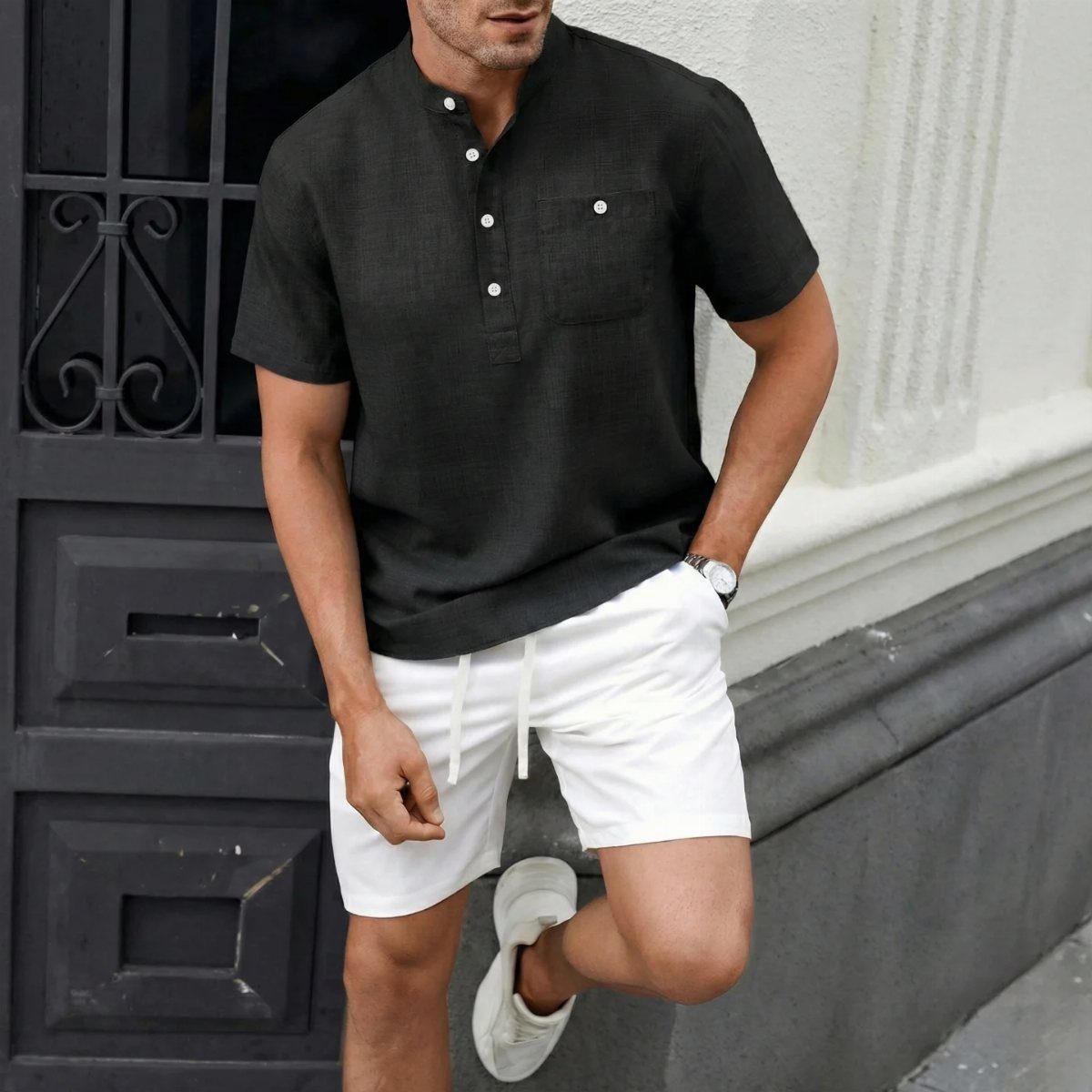 Man wearing a black shirt and white shorts posing against a white wall.