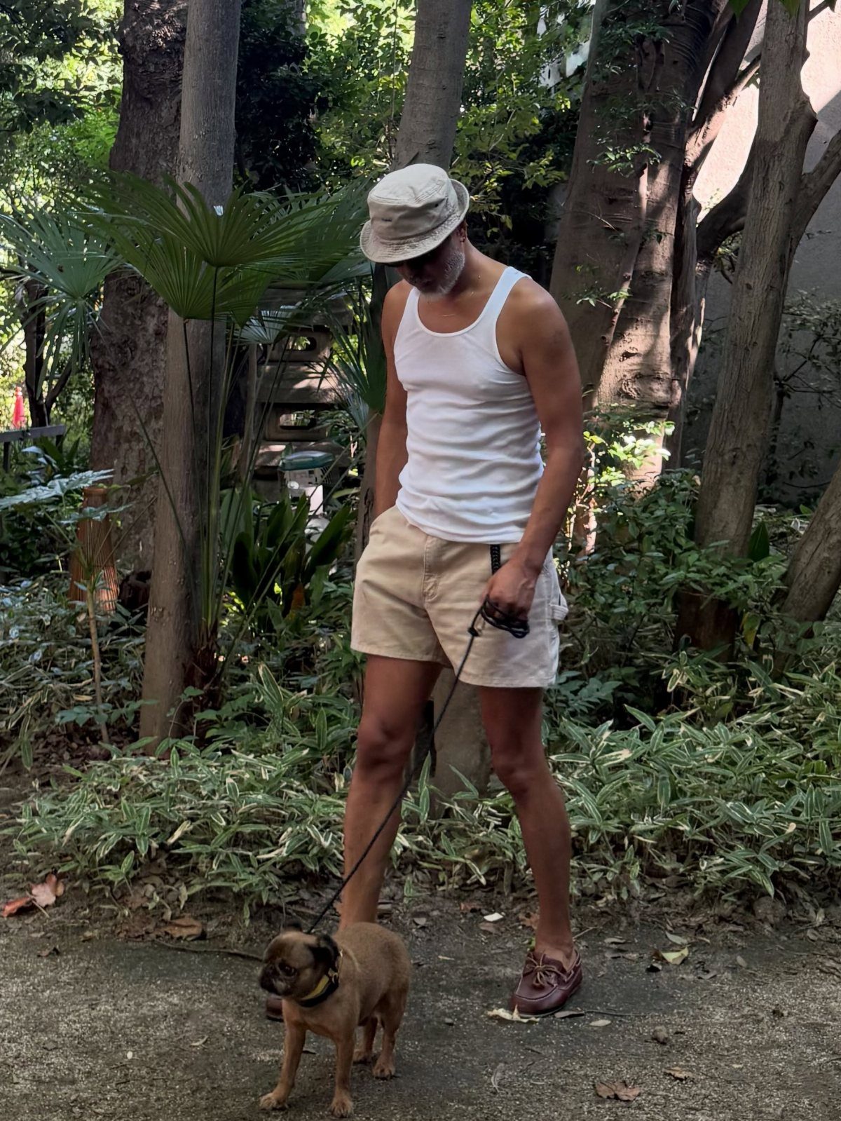 Man wearing white tank top and off-white shorts walking a small brown dog.