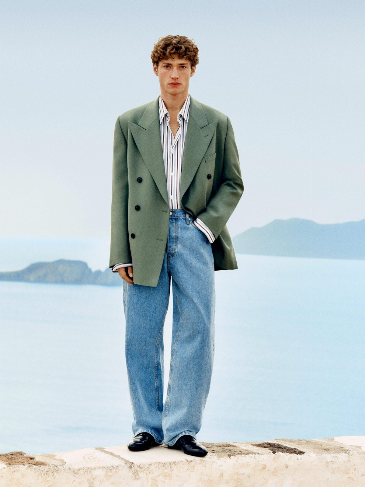 Man wearing a green blazer, striped shirt, and wide blue jeans by the ocean.