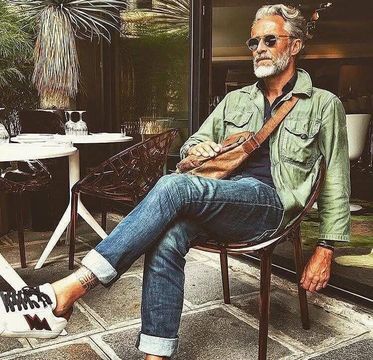 Mature man with grey beard wearing blue cuffed jeans and green shirt sitting outdoors.