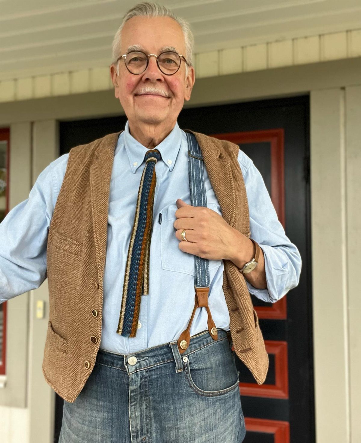 Older man wearing a brown vest, blue shirt, and blue jeans with suspenders.