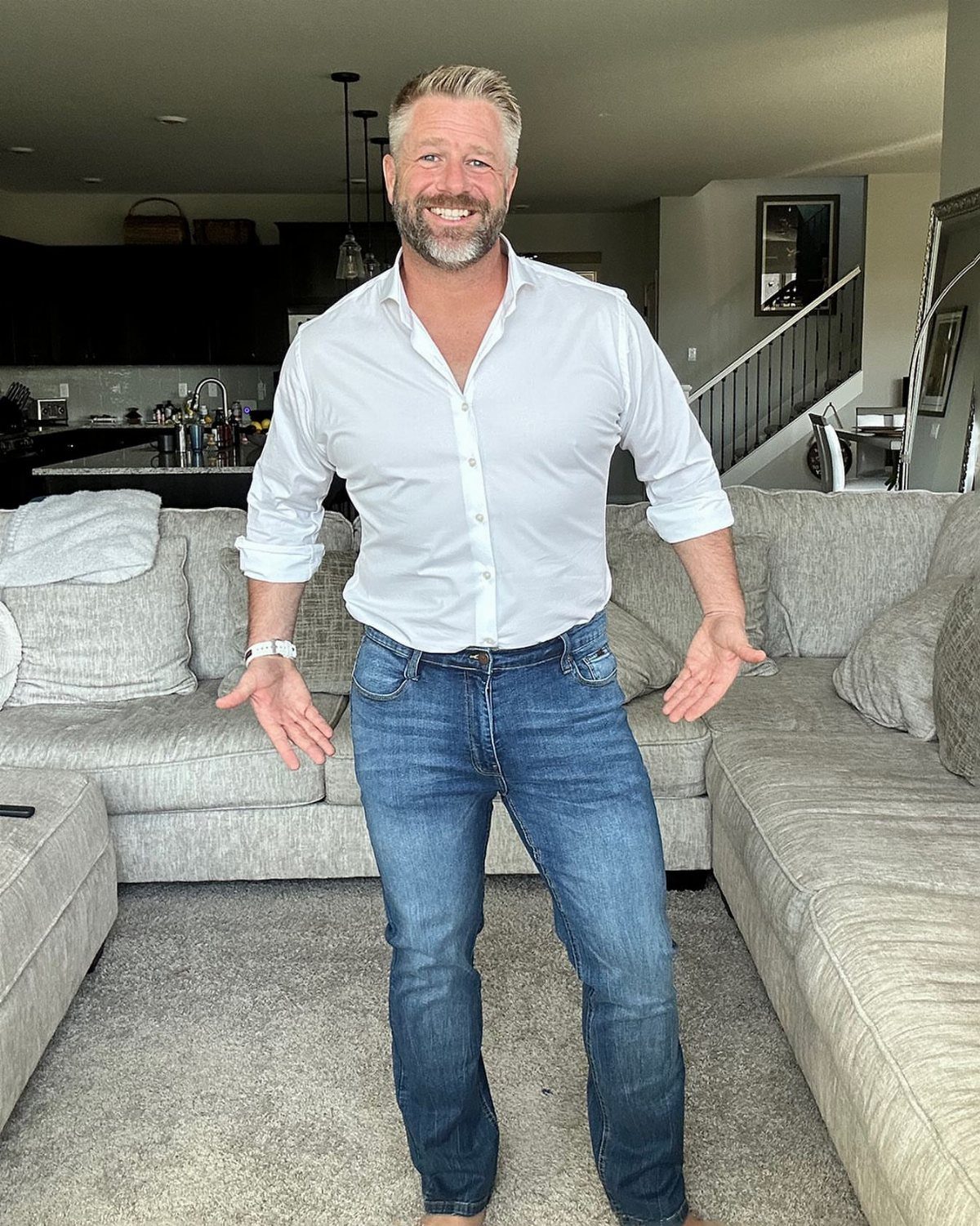 Middle-aged man wearing a white shirt and blue straight leg jeans in a living room.