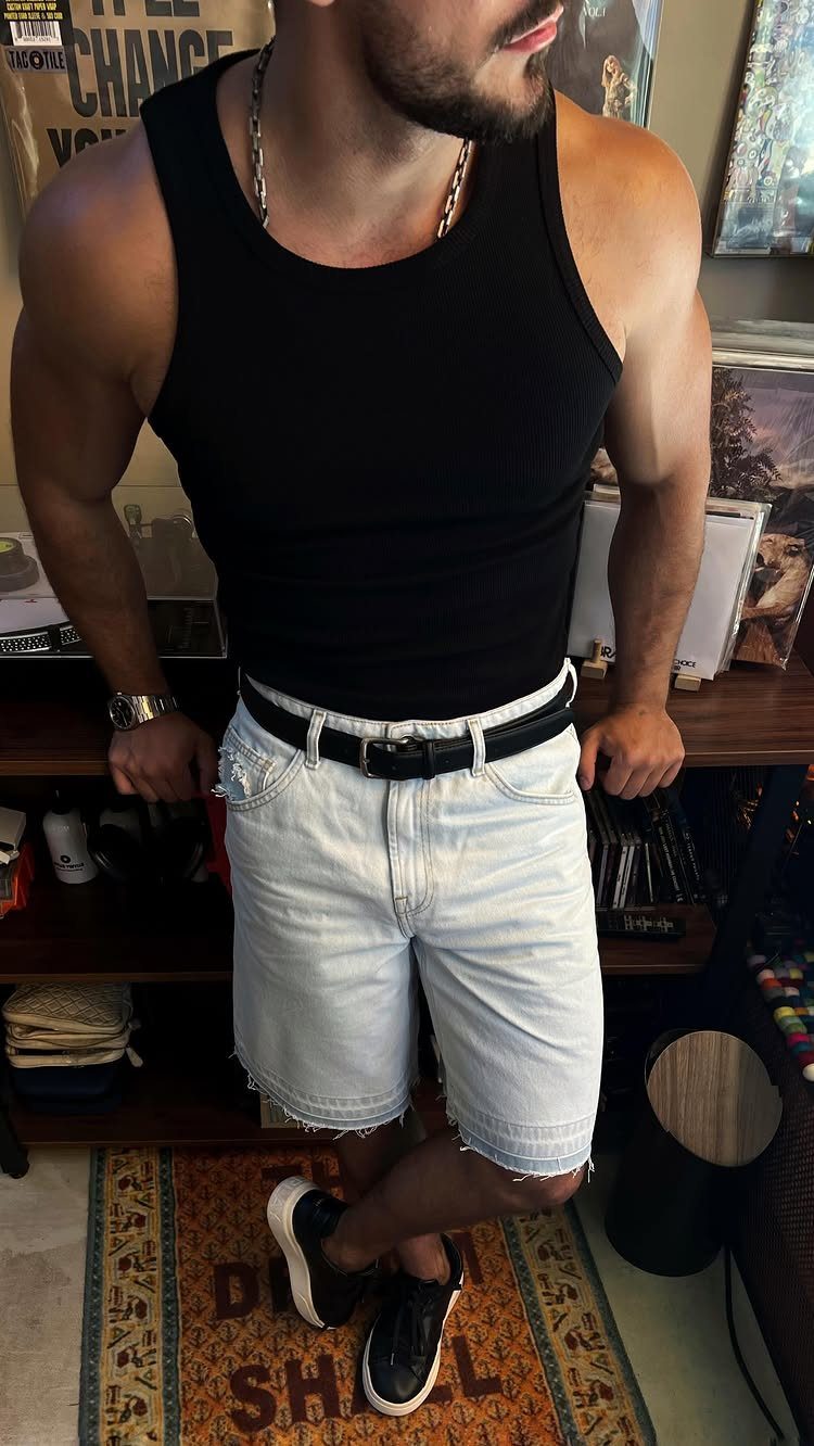 Man wearing a black tank top and light blue denim shorts standing indoors.