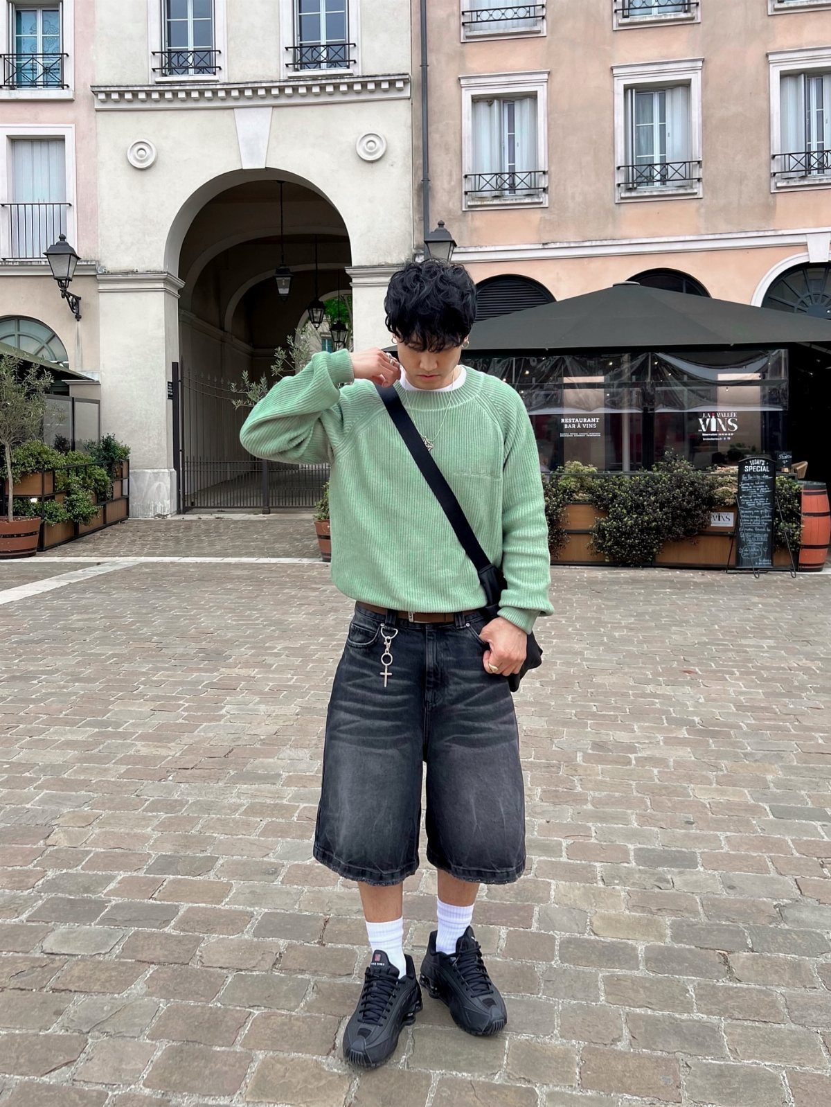 Person wearing a green sweater and wide-leg denim shorts on a cobblestone street.
