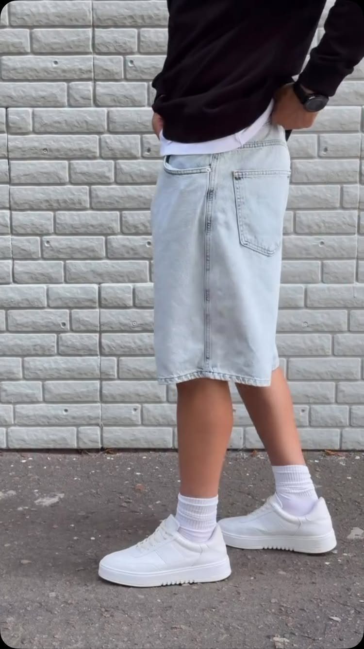 Person wearing light-wash denim shorts, black shirt, and white sneakers against brick wall.