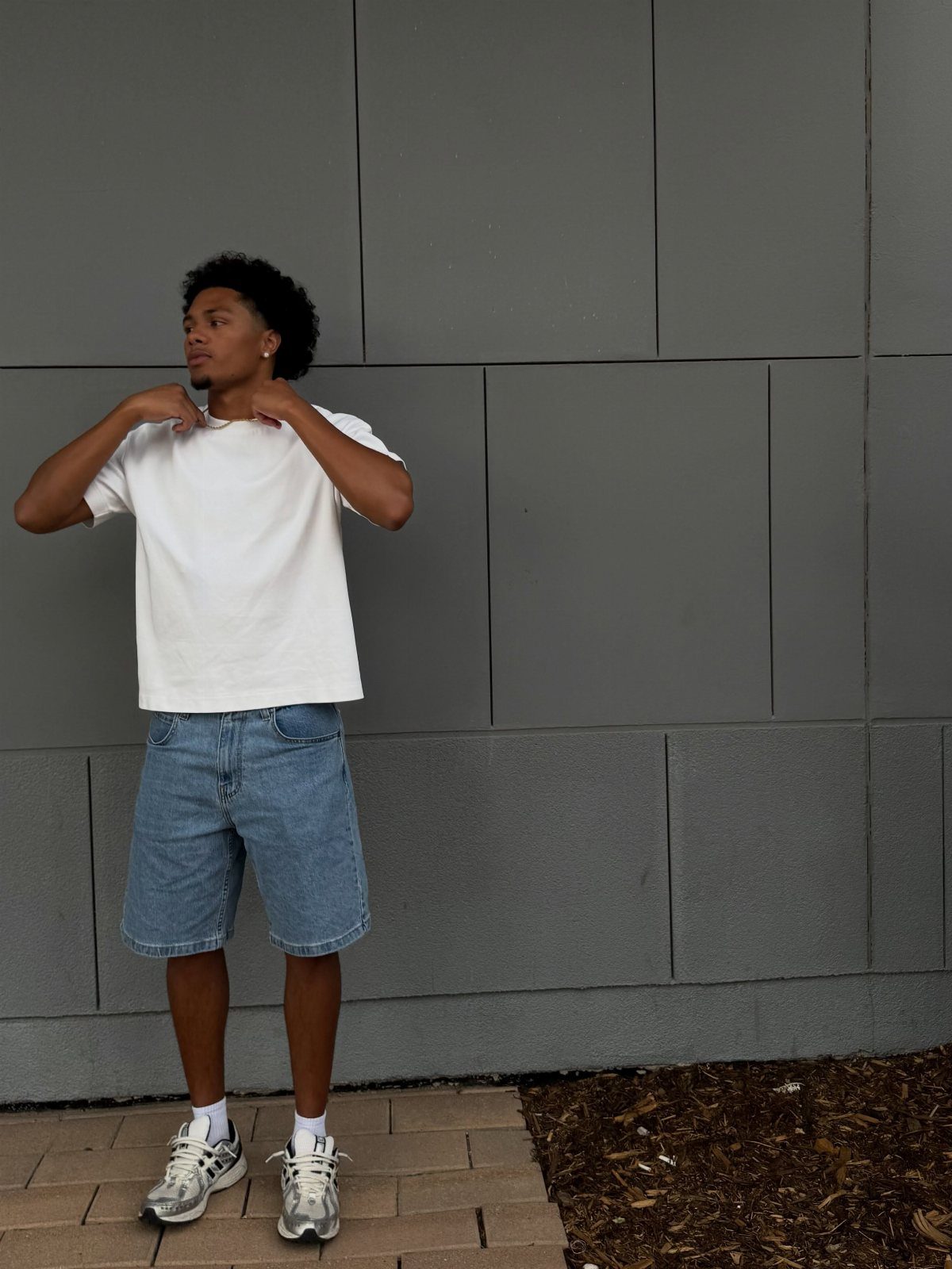 Man wearing a white t-shirt and blue denim shorts against a grey wall.
