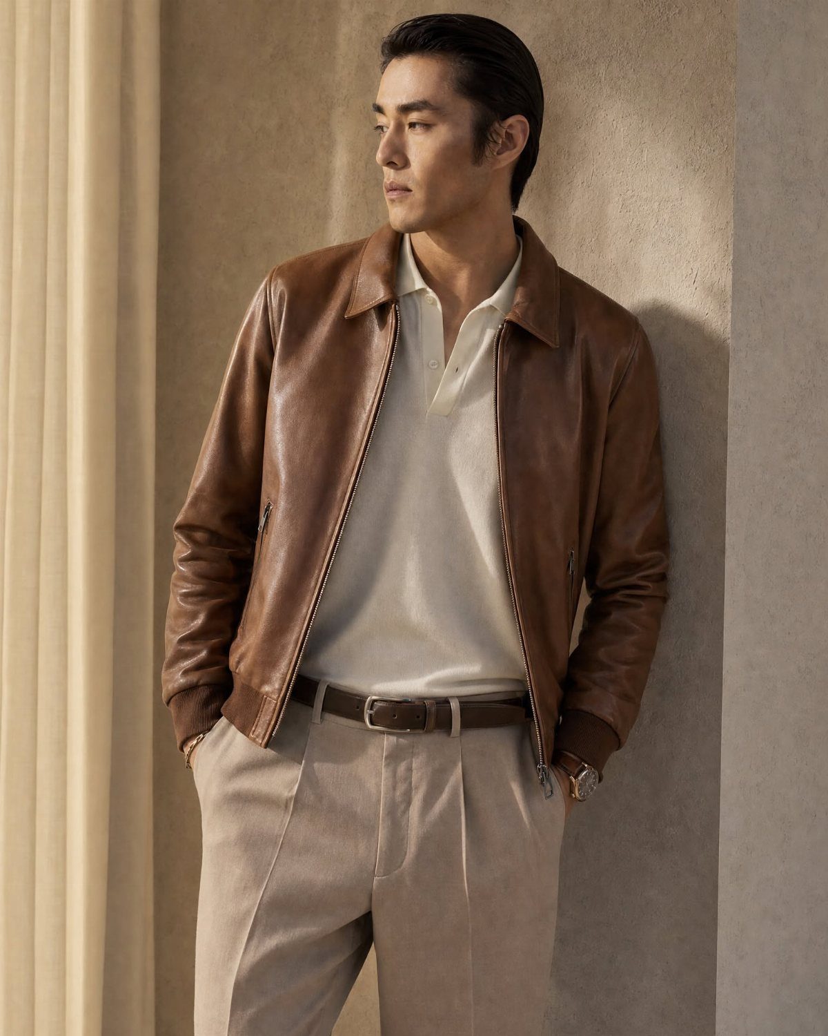 Man wearing a brown leather jacket, white polo, and khaki trousers leaning against wall.