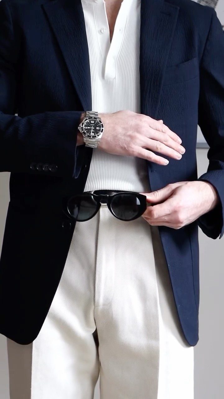 Man wearing a navy blazer, white shirt, and white linen pants holding sunglasses.