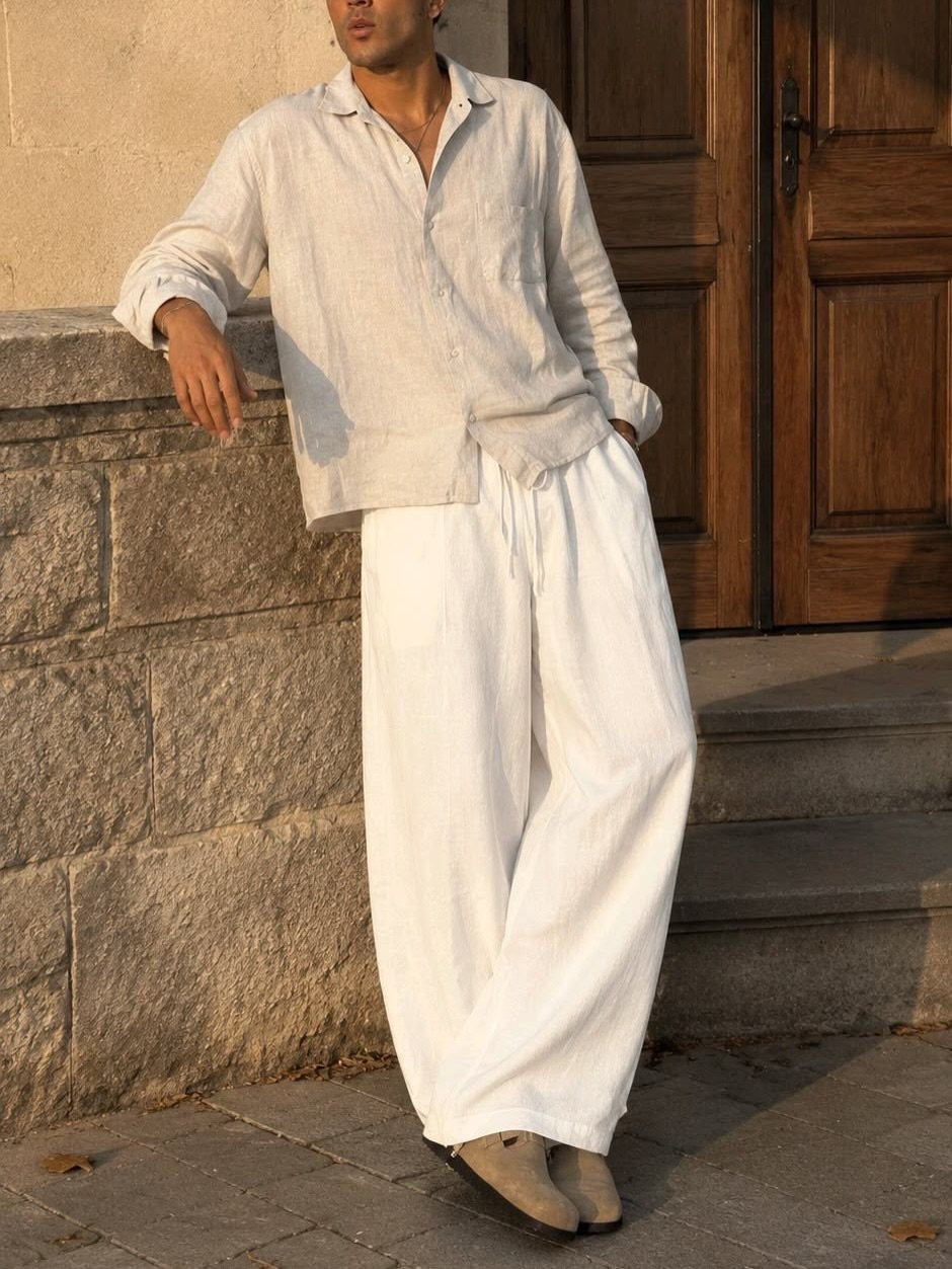 Man wearing a beige linen shirt and wide-leg white linen pants leaning on stone.