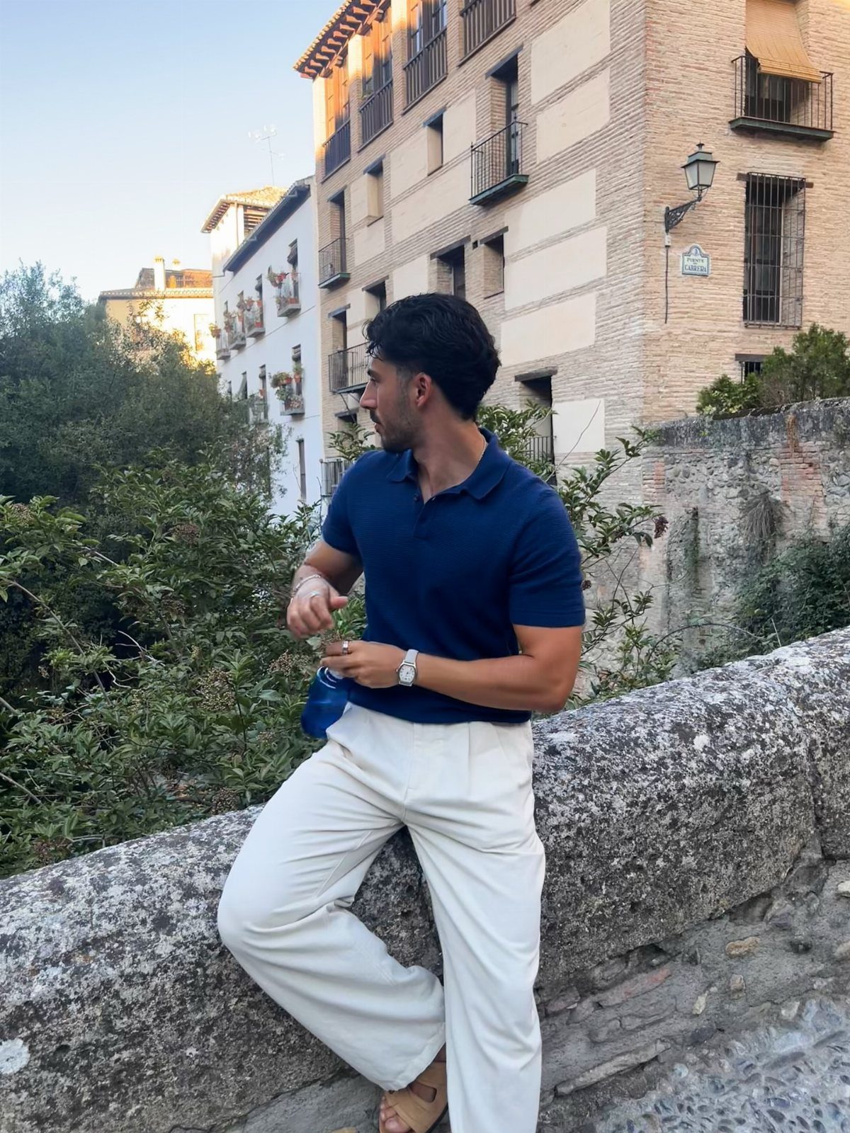 Man in navy blue polo shirt and white trousers leaning on stone wall