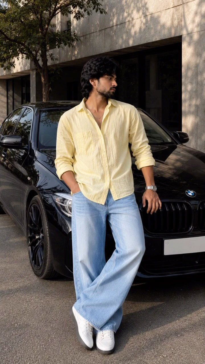 Man wearing a yellow linen shirt and light blue jeans leaning on a car